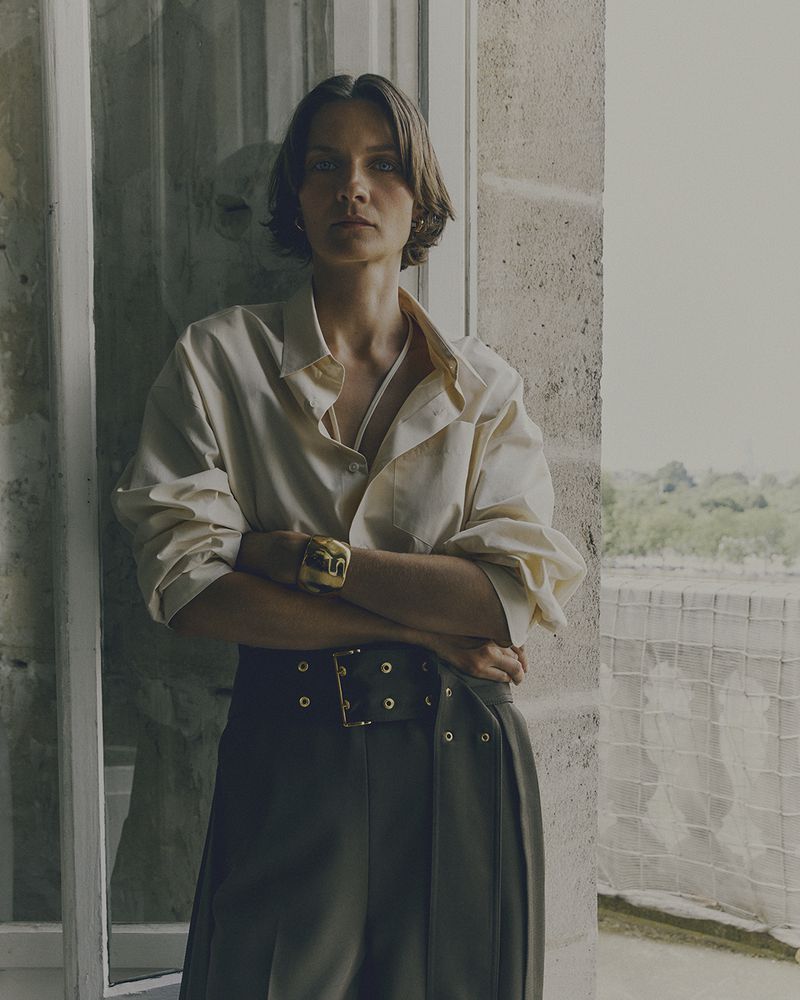 A woman in a white shirt and olive-green pants stands indoors with arms crossed, next to a window with muted natural light.