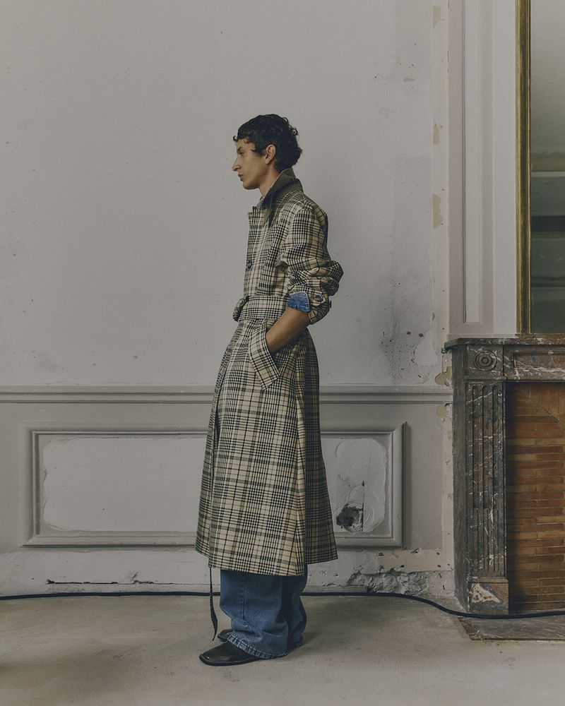 Side view of a person with curly dark hair wearing a long plaid coat, denim shirt, and jeans, standing indoors against a light gray wall.