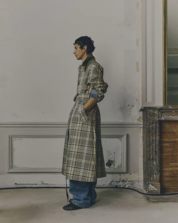 Side view of a person with curly dark hair wearing a long plaid coat, denim shirt, and jeans, standing indoors against a light gray wall.