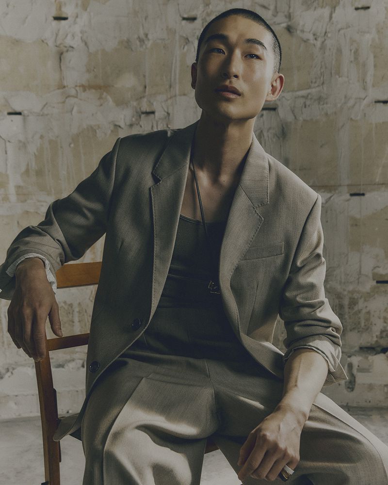 Asian male model in a tan suit sits on a wooden chair against a textured wall in a studio, looking at the camera with a neutral expression.