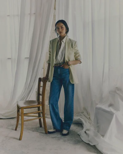 Model standing next to a wooden chair wearing an oversized blazer and blue denim jeans against white curtains.