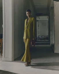 Model wearing an olive green blazer and flared trousers holding a handbag in a hallway