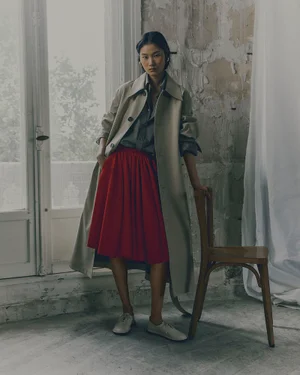 Model wearing a tan trench coat, grey shirt, and red skirt standing next to a wooden chair near a window