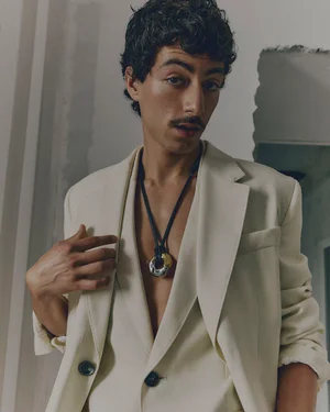 Man wearing a cream suit jacket and pendant necklace against an architectural wall