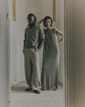 Two models posing in an unfinished interior space for Ami, wearing neutral tone apparel including wide-leg trousers and a sleeveless maxi dress.