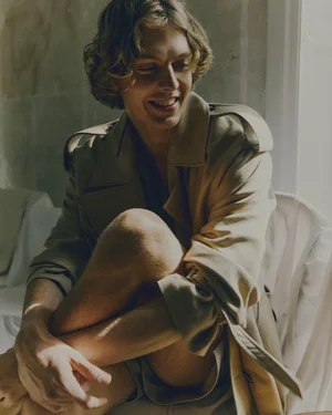 Portrait of a man with curly hair wearing a beige trench coat seated indoors near a window with natural light.