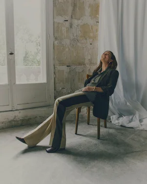 Smiling woman in dark blazer and two-tone trousers seated on a wooden chair against a distressed stone wall and white drape.