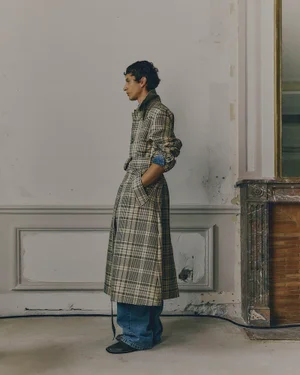 Model standing in profile wearing a long plaid trench coat and loose denim jeans beside a fireplace mantle.