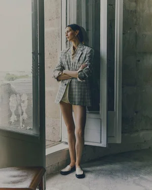 Model standing by a tall glass door wearing a plaid blazer and loafers for Ami