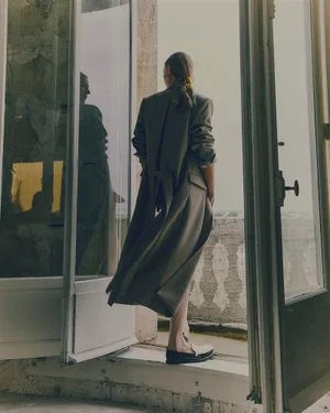 Model standing on a balcony threshold facing outward, wearing a long taupe coat and dark loafers.