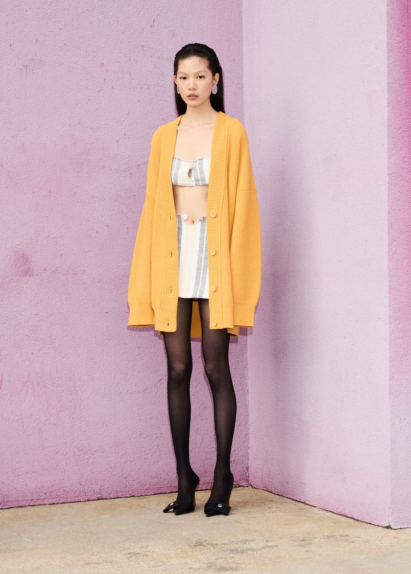 Full-length shot of an Asian fashion model wearing an orange cardigan, striped skirt and crop top, and black tights in a photo studio.