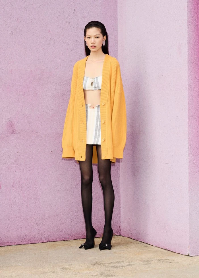 Model wearing a yellow oversized cardigan over a striped two-piece outfit against a pink wall.