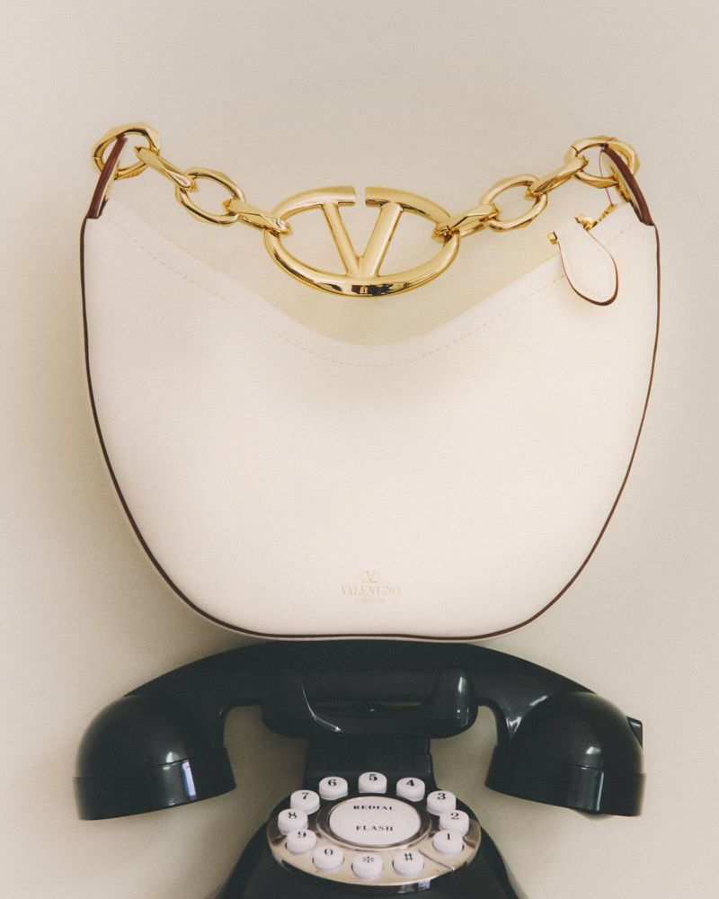 Valentino Garavani Loco small shoulder bag in ivory sitting on top of a black rotary telephone in a neutral setting.