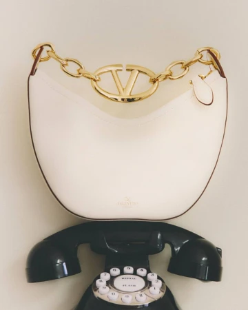 Valentino Garavani Loco small shoulder bag in ivory sitting on top of a black rotary telephone in a neutral setting.
