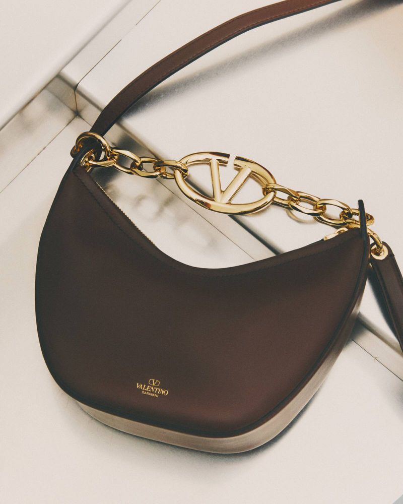Luxury Valentino Garavani hobo bag with VLogo chain, crafted in dark brown leather, suspended against a neutral gradient background.