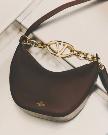 Luxury Valentino Garavani hobo bag with VLogo chain, crafted in dark brown leather, suspended against a neutral gradient background.