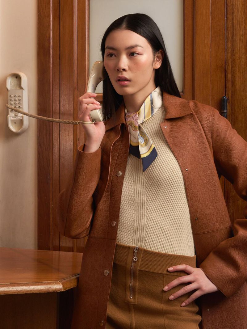 Elegant Asian woman in tan coat using vintage telephone in wood-paneled room, classic retro fashion style, indoor shot, soft lighting.