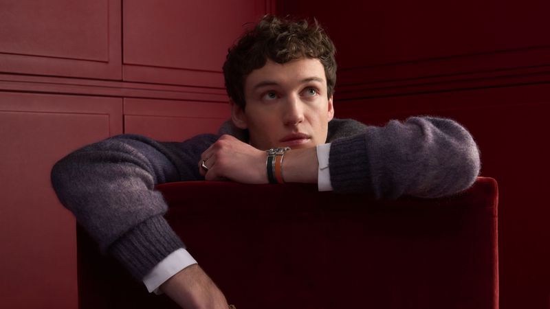 Young man with curly hair poses indoors with a grey sweater and bracelets. He leans on a red velvet surface in a portrait shot.