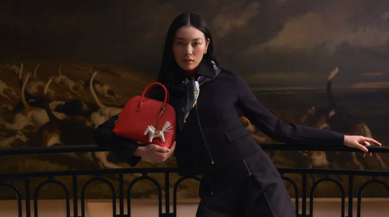 Woman posing in an indoor setting, wearing a black jacket, holding a red handbag with a white horse charm, and a painting in the background.