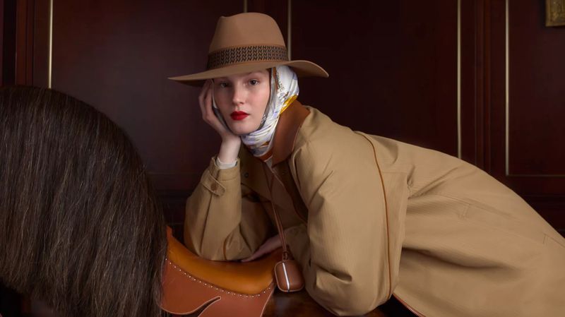 Adult female model wears tan coat and hat in front of partial horse and wood paneled wall. Dark indoor lighting and red lipstick.