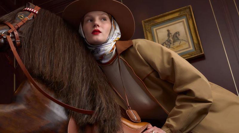 Adult female fashion model in a brown hat and jacket poses with a toy horse in an indoor setting with an ornate painting on the wall.