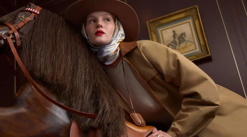 Adult female fashion model in a brown hat and jacket poses with a toy horse in an indoor setting with an ornate painting on the wall.