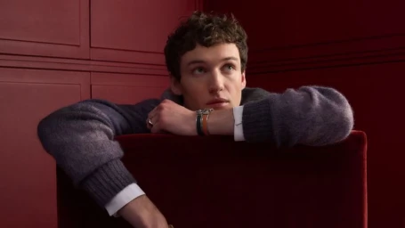 Close-up portrait of a male model in a grey knit sweater leaning over a dark red velvet chair inside a matching crimson room