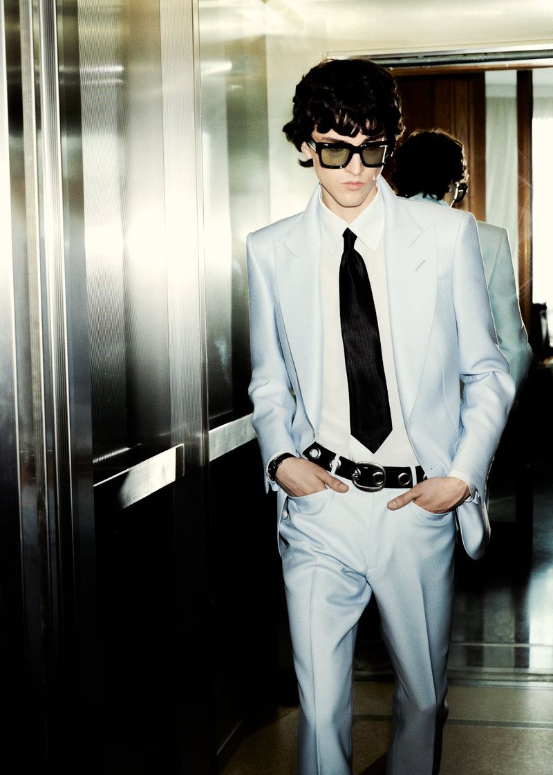 A man poses in a light blue suit with a black tie, wearing dark sunglasses, standing indoors with a mirrored wall in the background.