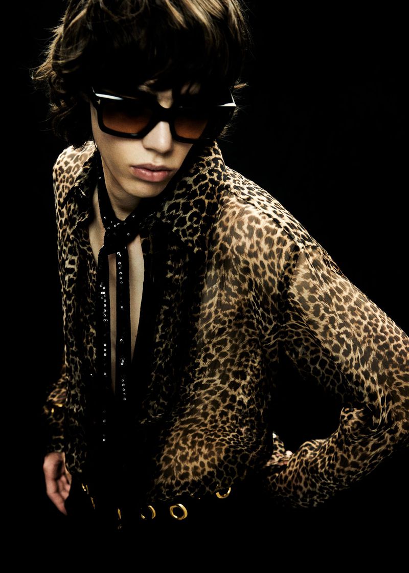 A person poses in a cheetah-print shirt and sunglasses against a black background, fashionably styled in a studio setting.