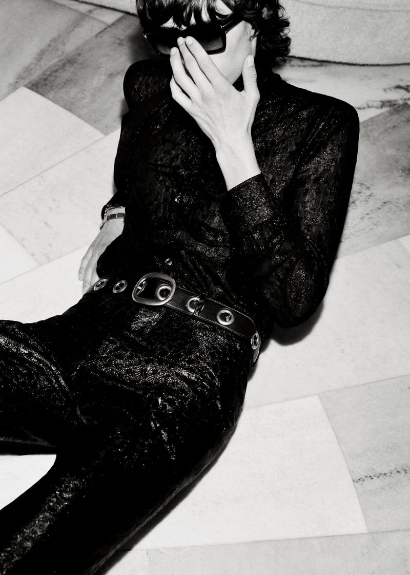 Person in a black, textured outfit and sunglasses reclines on a marble floor in a high-contrast black and white portrait, fashion.
