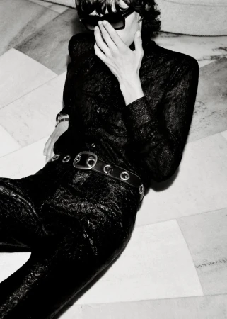 Person in a black, textured outfit and sunglasses reclines on a marble floor in a high-contrast black and white portrait, fashion.