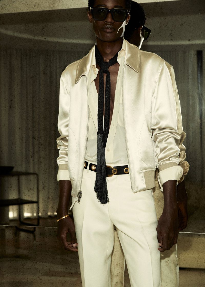 A stylish studio shot presents a man in a satin bomber jacket, dress shirt, and pants, accessorized with sunglasses and a black tassel tie.