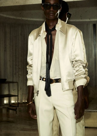A stylish studio shot presents a man in a satin bomber jacket, dress shirt, and pants, accessorized with sunglasses and a black tassel tie.