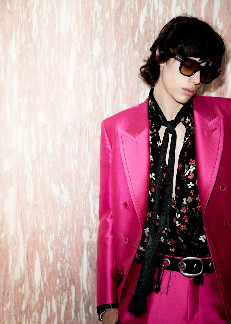 Young adult with dark hair wearing sunglasses, a floral shirt, and a hot pink suit jacket and pants poses against a textured wall.