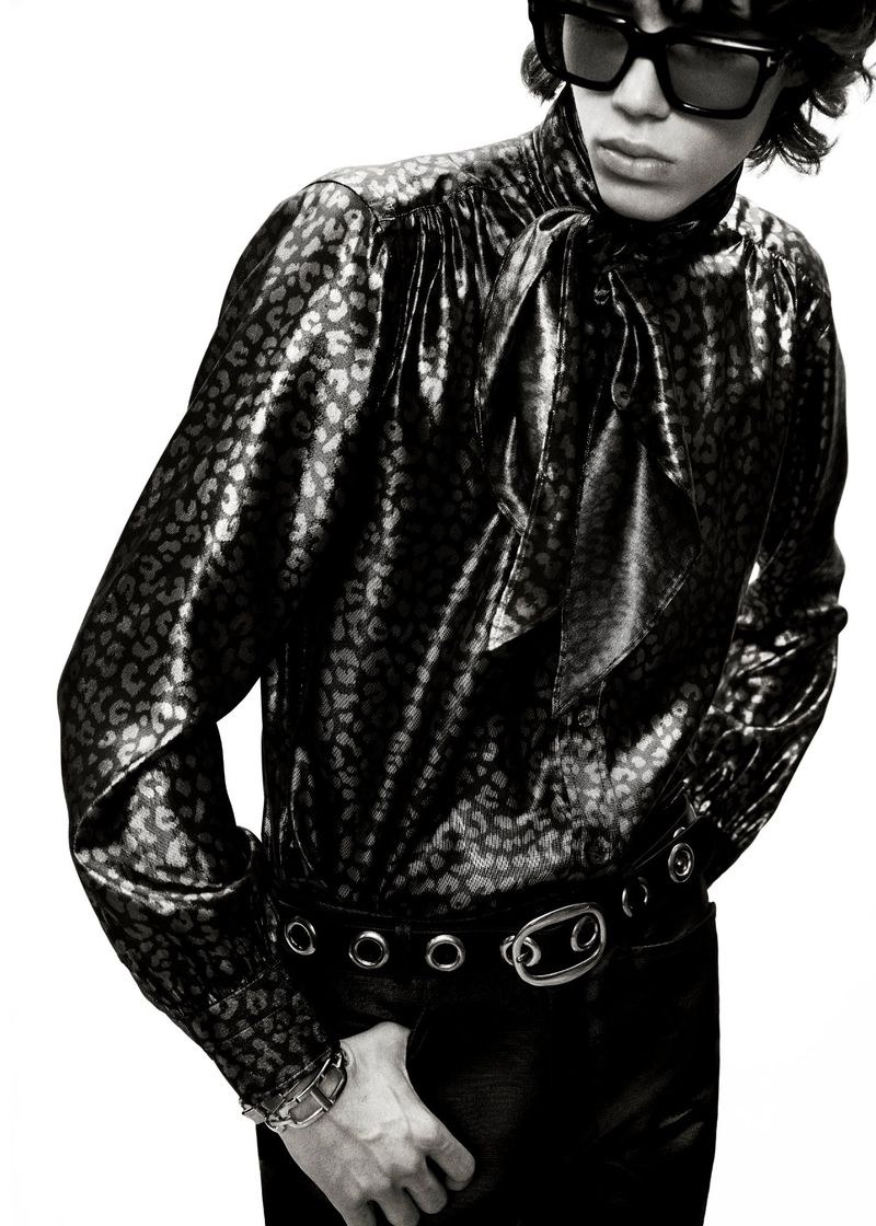 Black and white fashion portrait of a person with dark hair and sunglasses wearing a patterned shirt with a pussy bow and leather pants.