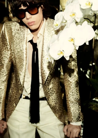 A stylish young man in a gold leopard print jacket and sunglasses poses near white orchids, showcasing a high-fashion editorial look.