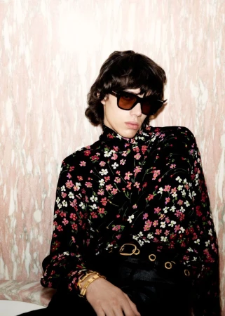 Young adult with sunglasses wears a black floral shirt, accessorized with gold bracelets and belt against a pink marble backdrop.