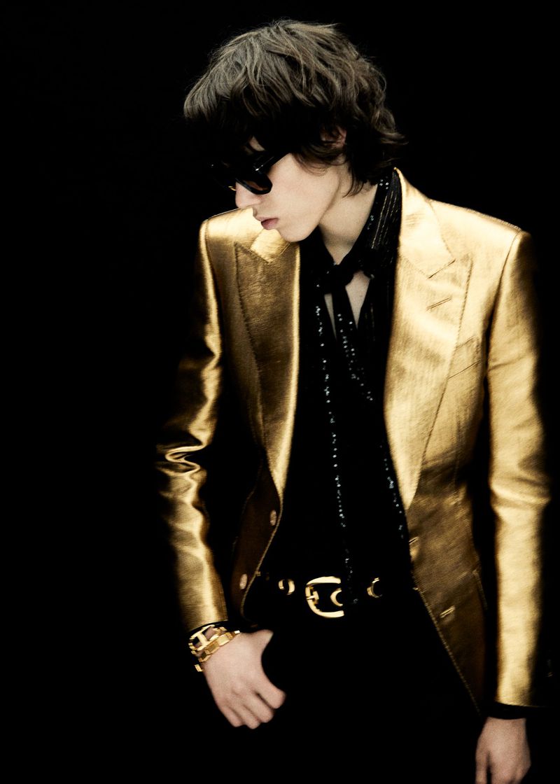 Stylish adult man in a golden blazer and dark sunglasses posing against a dark background in a fashion style photograph.