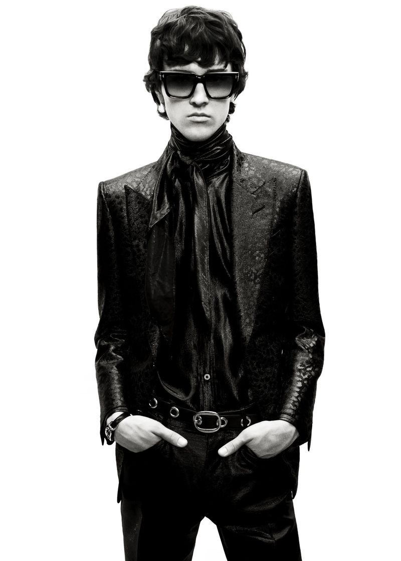 Monochrome portrait of a person with dark wavy hair, wearing a blazer, black shirt, sunglasses, and jewelry against a bright background.