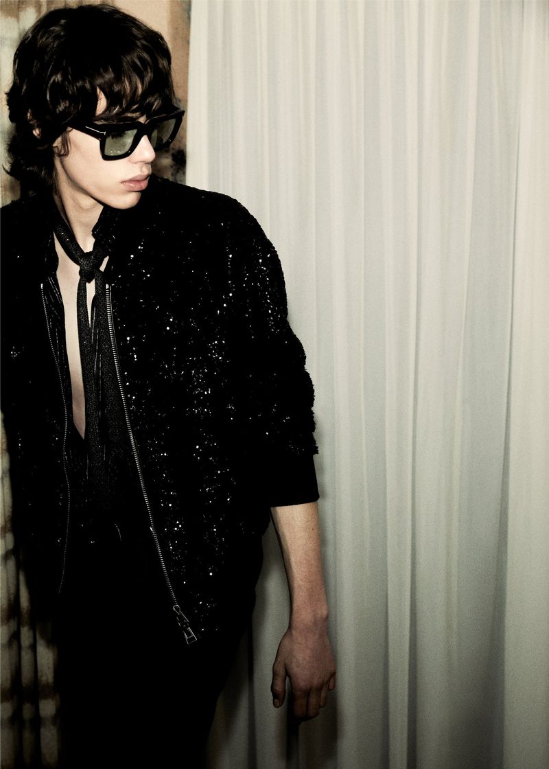 Adult male in a dark setting wearing a black sequin jacket and sunglasses, looking off to the side with a curtain behind him.