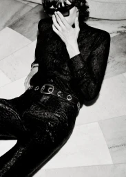 Model reclining on a marble floor wearing sunglasses and a textured black outfit with a wide grommet belt.