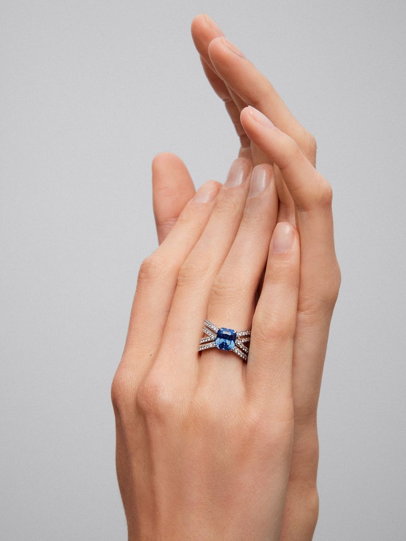 Close-up of a hand wearing a blue sapphire ring with diamond accents against a neutral background, emphasizing jewelry design and detail.