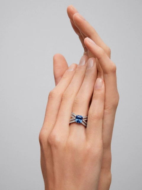 Close-up of a hand wearing a blue sapphire ring with diamond accents against a neutral background, emphasizing jewelry design and detail.