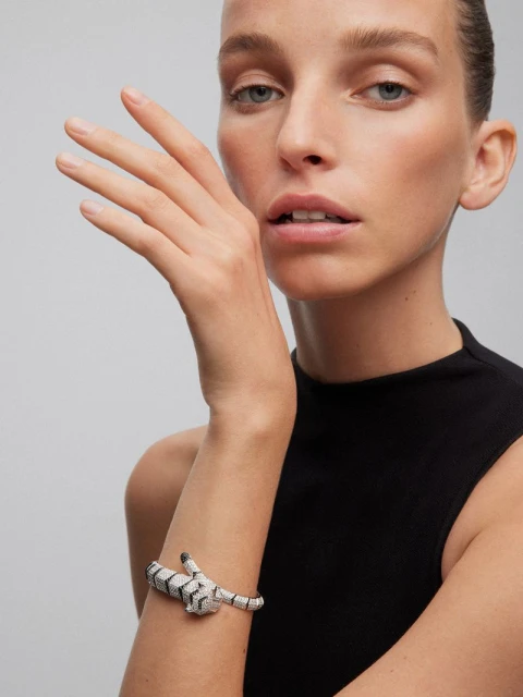 Close-up of a woman with fair skin and light hair wearing a black top and silver and black bracelet in a bright indoor setting.