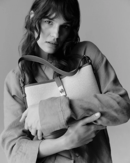 Black and white portrait of a brunette woman in a shirt holding a woven handbag with leather straps indoors.