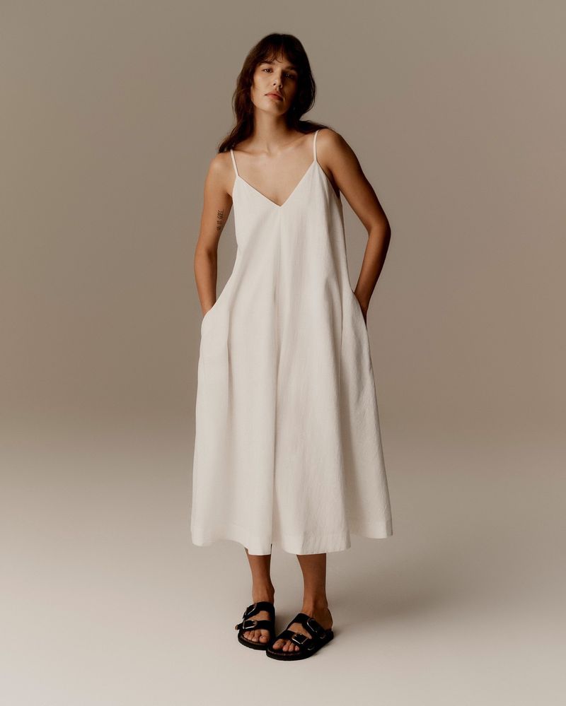 Adult woman in a studio wearing a cream V-neck dress with thin straps and flat black slide sandals poses with hands in her pockets.
