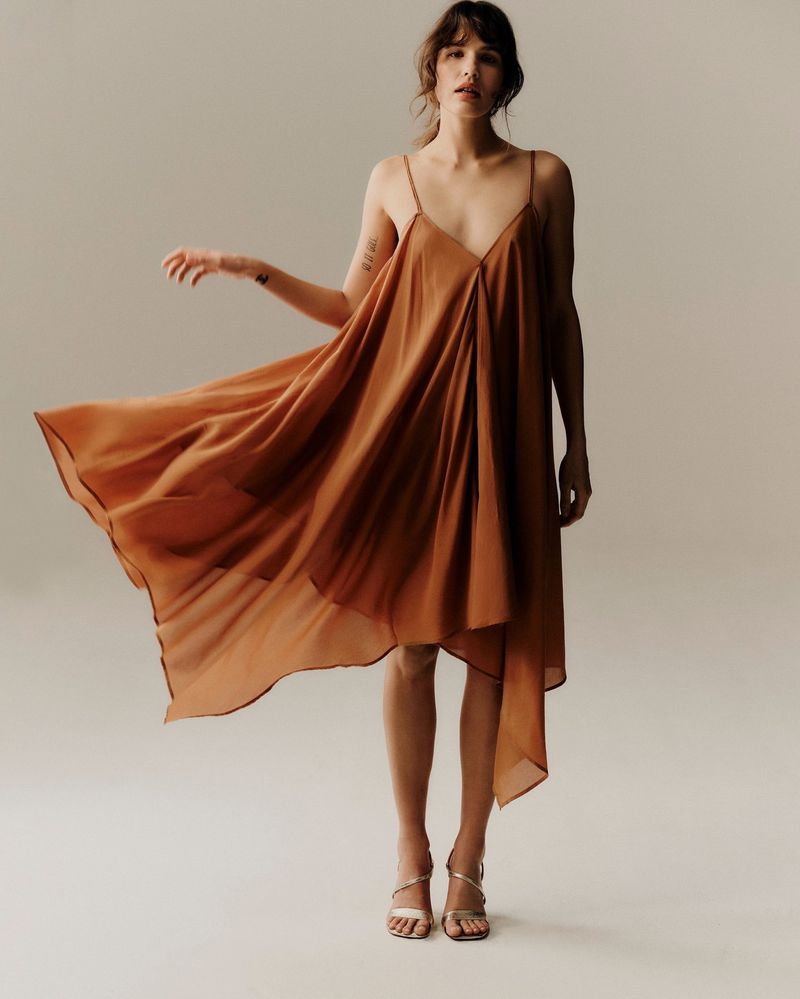 A fashionable woman in a flowing copper-colored dress with spaghetti straps, posing in a photo studio against a neutral backdrop.