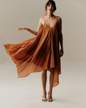 A fashionable woman in a flowing copper-colored dress with spaghetti straps, posing in a photo studio against a neutral backdrop.