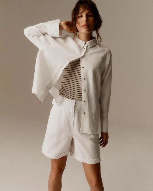 Adult woman poses in a white linen shirt and shorts inside a photo studio, showcasing a casual fashion style with natural lighting.