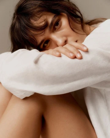 Portrait of a brunette woman with her head resting on her arms, wearing a white top, posing indoors against a neutral backdrop in soft lighting.